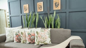 Un canapé gris agrémenté de coussins à motifs floraux et de grandes plantes vertes, le tout placé devant un mur sombre orné d’œuvres d’art encadrées.