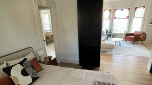 A spacious bedroom with a large bed, a black door, and a view of a dining area with a red chair and a plant. - Modern Comfort in the Heart of Cherry Hill (Grand Rapids)