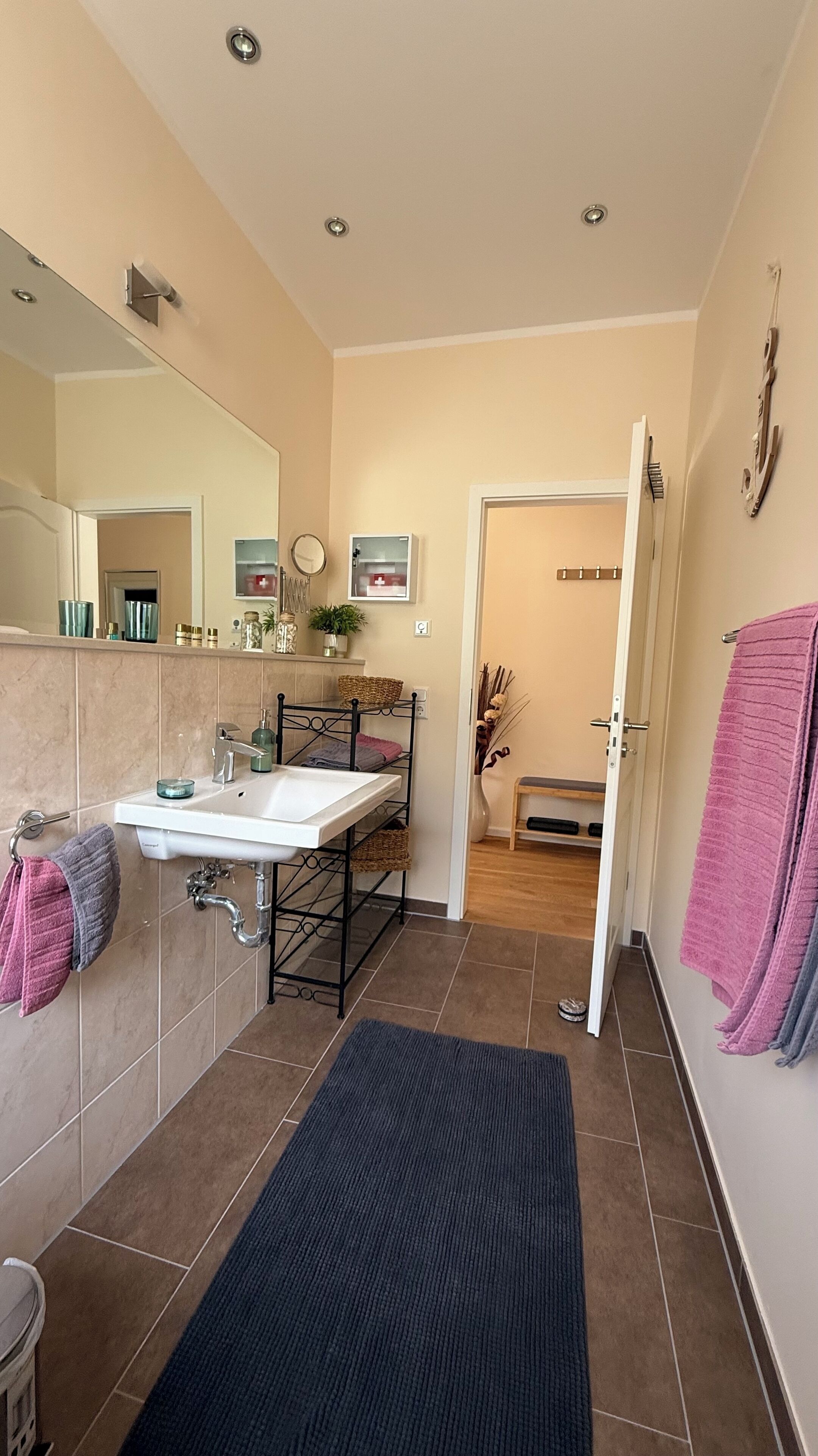 A modern bathroom with a white sink, mirror, and tiled floor. There is a black rug in the center, and towels are hung on the wall.