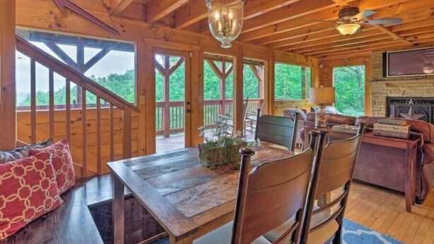 A wooden cabin interior with a dining table, chairs, a fireplace, and large windows offering a view of the outdoors. - Awesome Private Mountain views Resort Pool &HotTub (Sevierville)