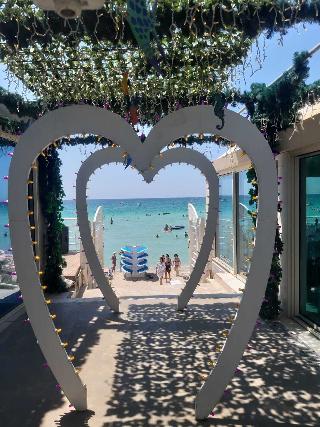 Un arco en forma de corazón que da paso a una playa con agua azul cristalina y gente nadando.