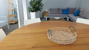 Una mesa de madera con una cesta tejida en el centro, un sofá blanco con cojines azules y una planta en maceta al fondo.