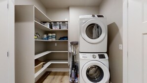 Un lavadero con lavadora y secadora apiladas, estantes con productos de limpieza y un aspirador.