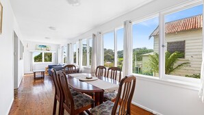 A dining area with a wooden table and chairs, large windows with curtains, and a view of greenery and buildings outside. - The Narooma Escape (Narooma)