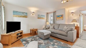 A living room with a flat-screen TV, grey sofa, ottoman, wooden TV stand, and a patterned rug. - Two Hoots - Peaceful Cottage Rural Views (Pembroke)
