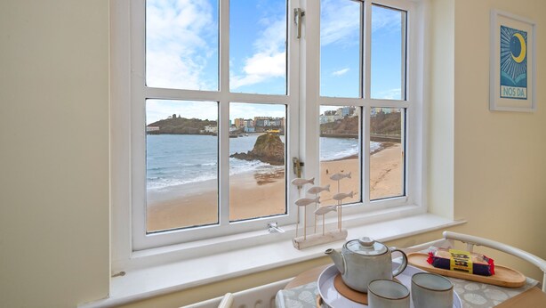 A room with a large window overlooking a beach, a tea set on a tray, and a framed picture on the wall. - 5 Sunny Cove - Direct Beach Access Sea Views (Tenby)