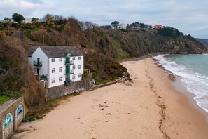 A coastal building with a white facade and green accents, situated on a sandy beach with a rocky cliff in the background. - 5 Sunny Cove - Direct Beach Access Sea Views (Tenby)