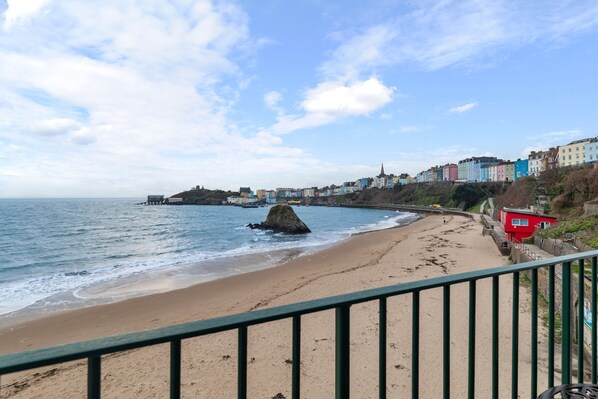 A coastal view with a sandy beach, a rocky outcrop, and a row of colorful buildings along the shore. - 5 Sunny Cove - Direct Beach Access Sea Views (Tenby)