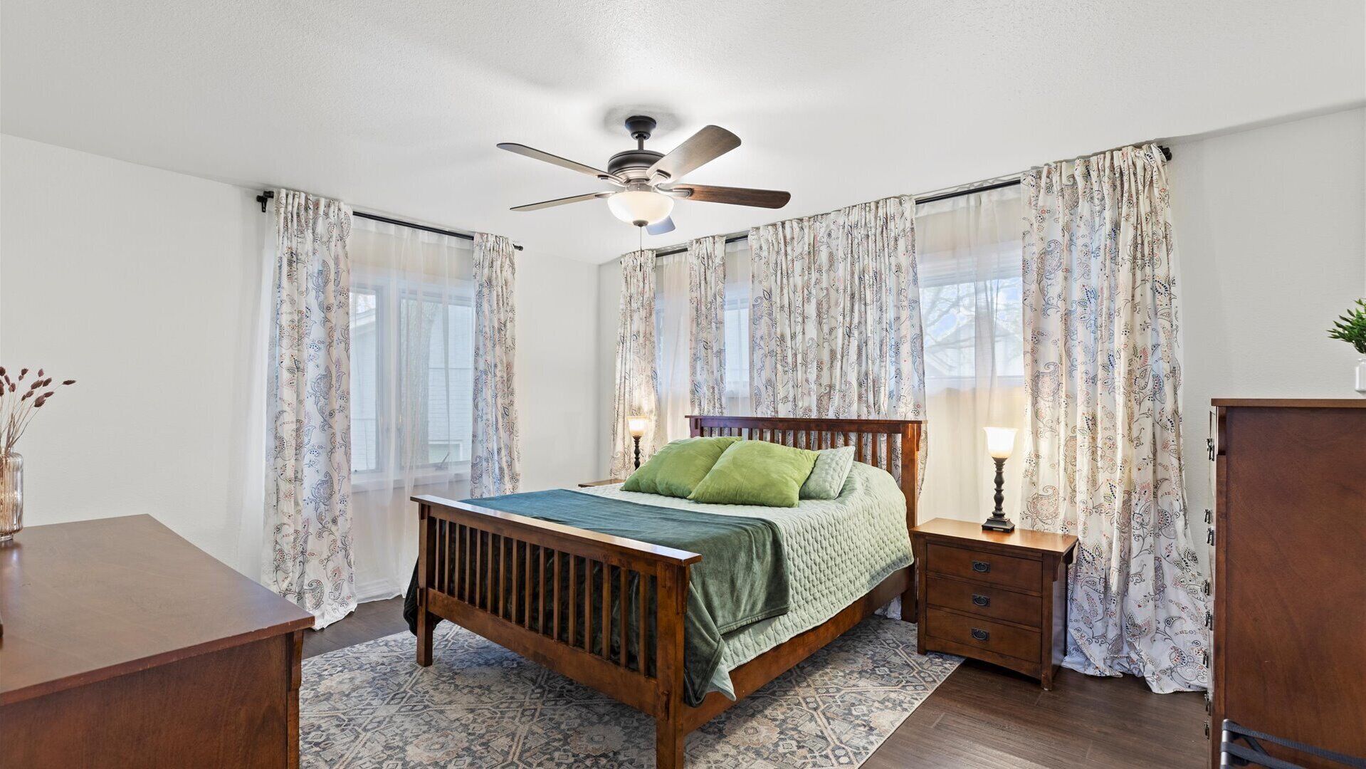 Une chambre à coucher avec un lit en bois, une table de chevet avec une lampe, des rideaux à motifs et un ventilateur de plafond.