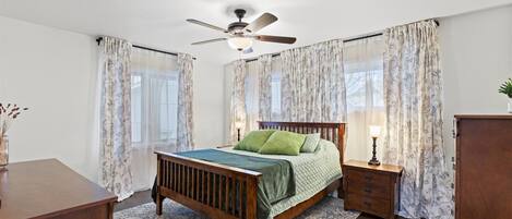 Une chambre à coucher avec un lit en bois, une table de chevet avec une lampe, des rideaux à motifs et un ventilateur de plafond.