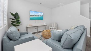 Una sala de estar moderna con un sofá gris, una mesa de centro blanca, un reposapiés de mimbre, un televisor de pantalla plana grande en la pared y una planta en maceta.