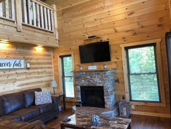 L’intérieur chaleureux d’un chalet avec une cheminée en pierre, un canapé en cuir et un téléviseur fixé au-dessus de la cheminée.