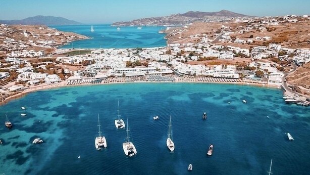 An aerial view of a coastal town with white buildings, a marina with several sailboats, and a clear blue sea.