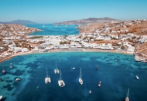 An aerial view of a coastal town with white buildings, a marina with several sailboats, and a clear blue sea.