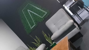 A modern lounge area with a green wall logo, cushioned seating, and a small wooden table. - HOTEL CAPITAL (Bogotá)