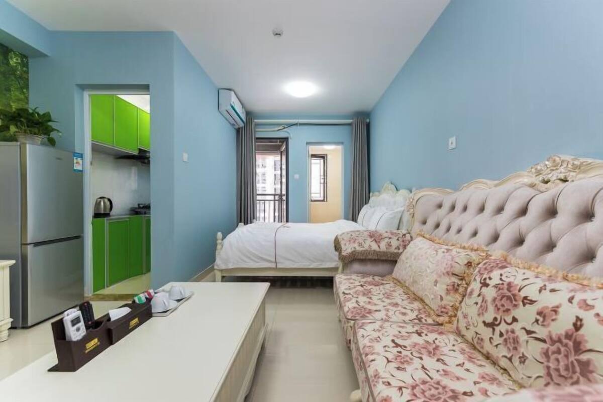 A compact room with a bed, a sofa, a small table, a kitchenette, and a bathroom door.