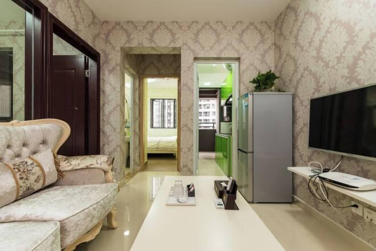 A living room with a sofa, a flat-screen TV, a refrigerator, and a kitchen area visible through an open doorway.