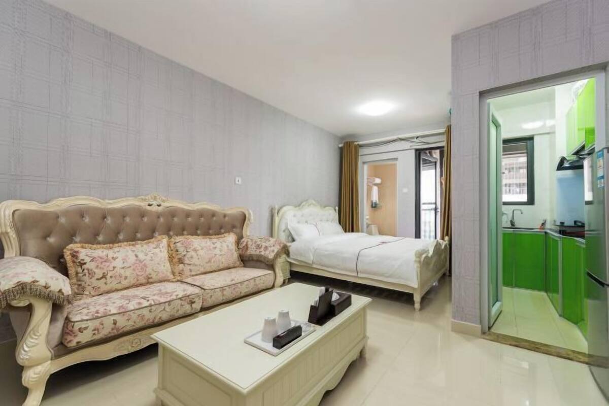 A room with a floral sofa, a bed, a coffee table, and a kitchenette.