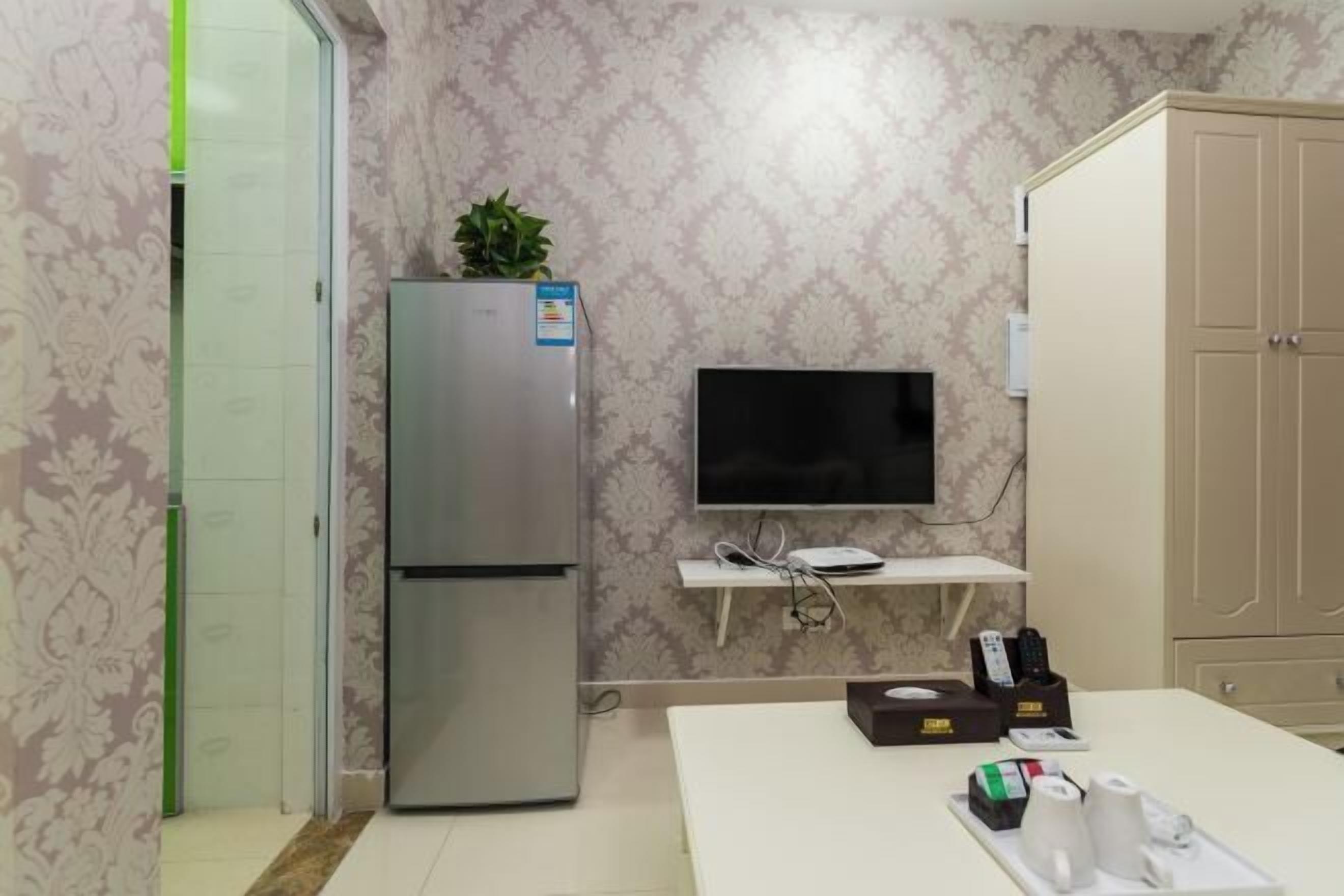 A kitchenette with a refrigerator, a TV, a small shelf with a plant, and a table with a tray of items.