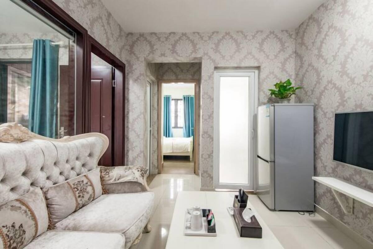 A hotel room with a sofa, a television, a refrigerator, and a hallway leading to a bedroom.