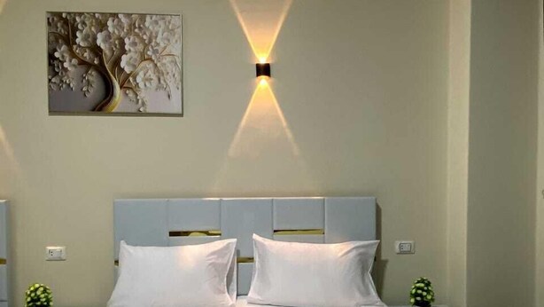 A hotel room with a large bed, two bedside tables, a wall-mounted light fixture, and a floral artwork on the wall. - Sky light hotel (Cairo)