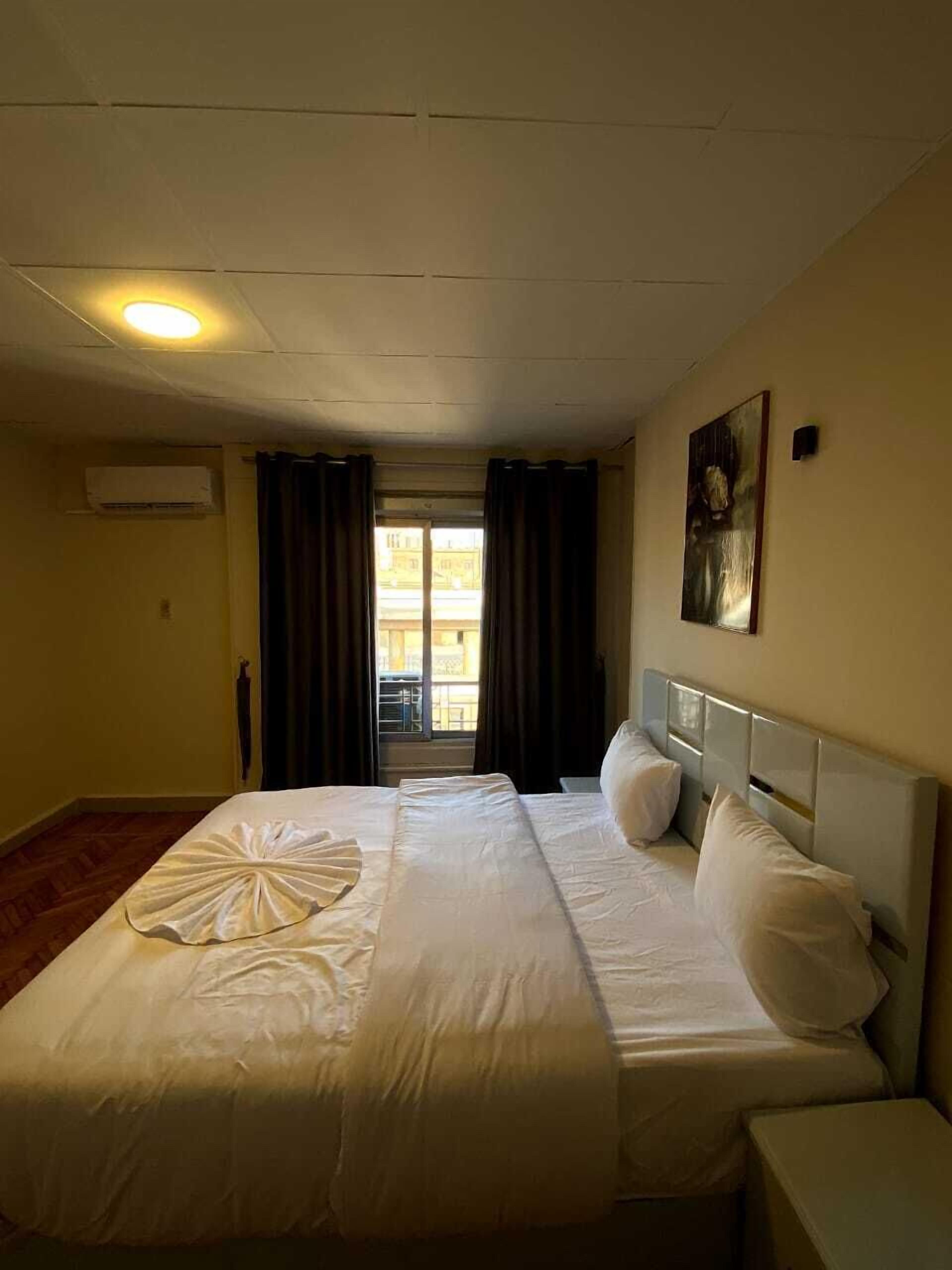 A hotel room with a bed, a nightstand, a painting on the wall, and a window with curtains.