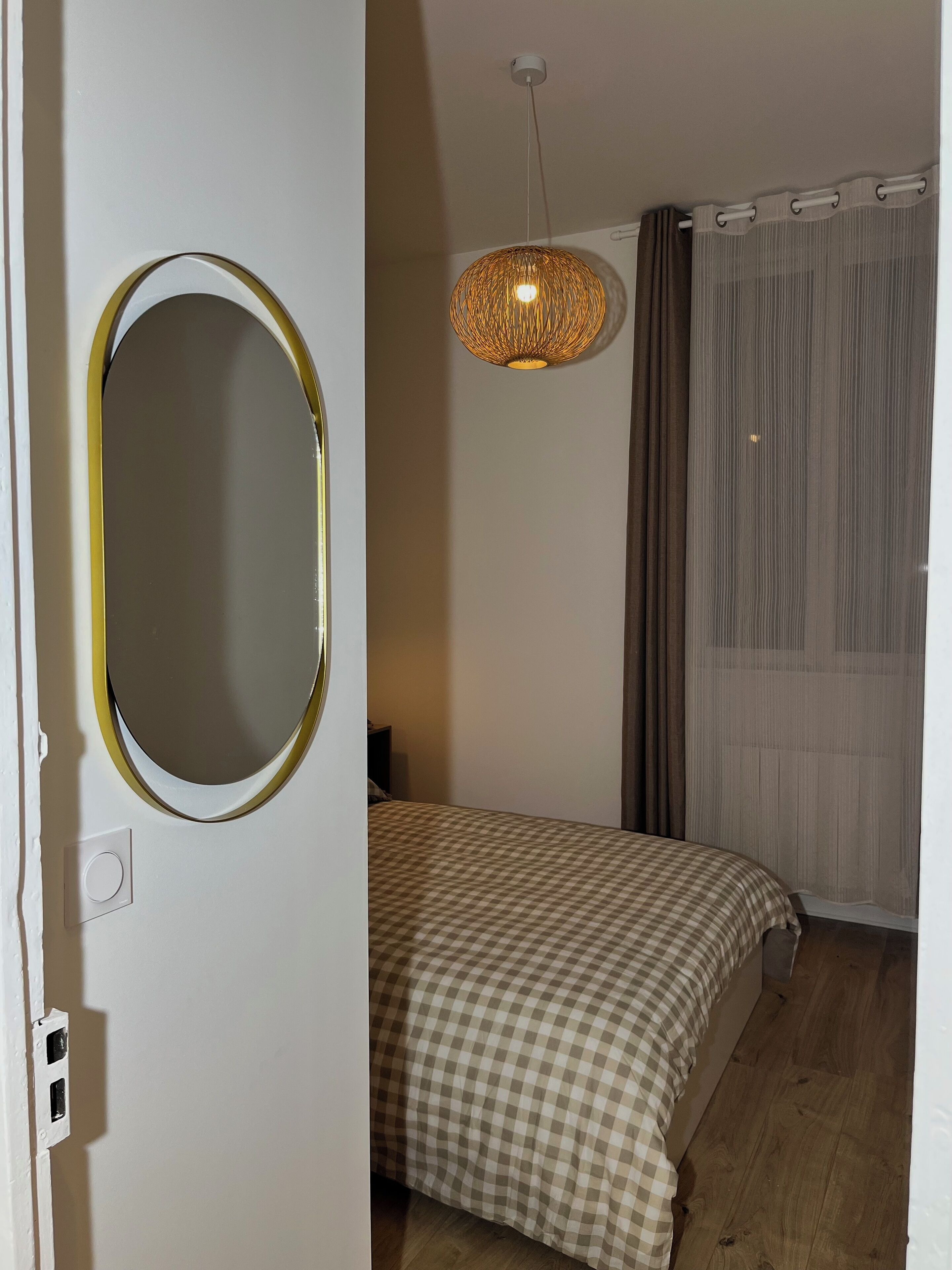 A bedroom with a bed, a round mirror, a hanging light fixture, and a window with curtains.