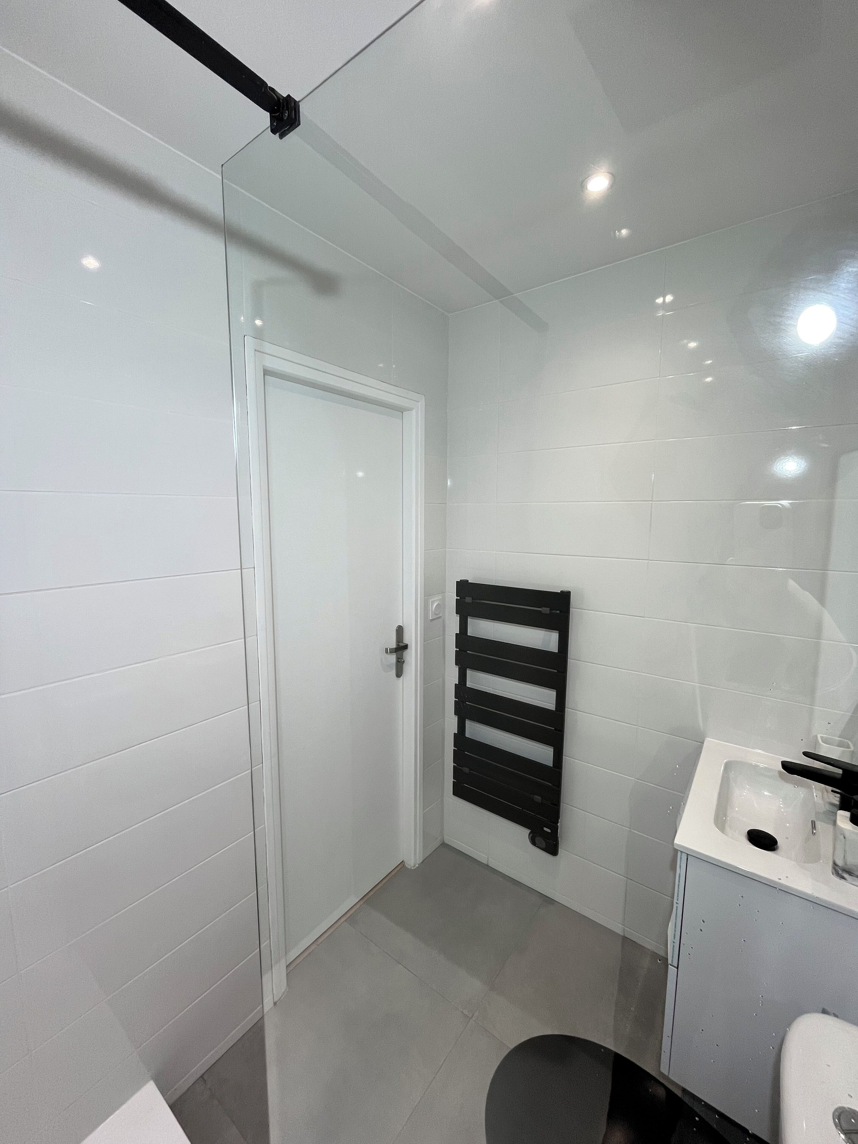 Un baño moderno con paredes de azulejos blancos, cabina de ducha de vidrio, toallero negro y lavamanos blanco con espejo.