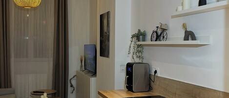 A modern kitchen with a built-in oven, a sink, and a coffee machine. There is a dining area with a table and chairs, and a living room with a sofa and a TV.
