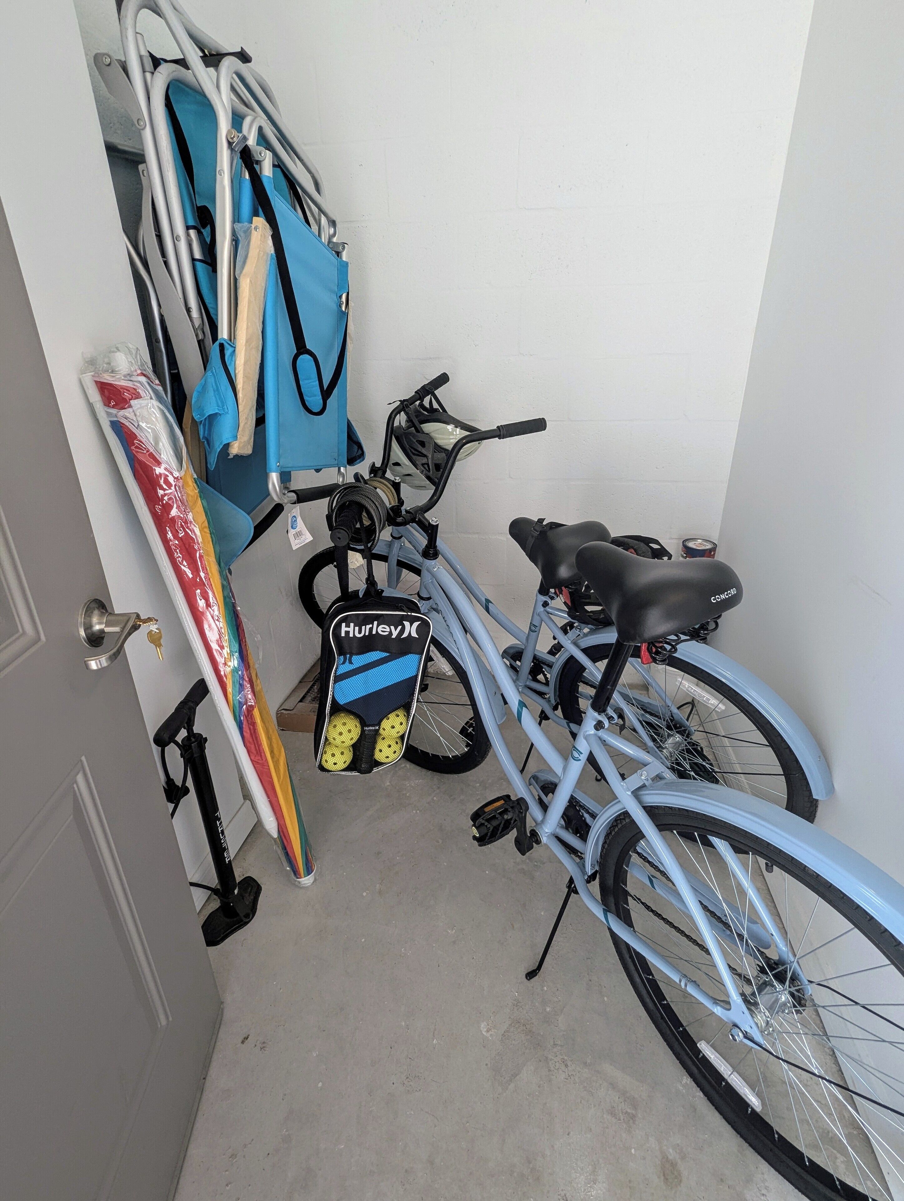 A bicycle with a Hurley brand seat, a blue bag, and a surfboard leaning against a wall.