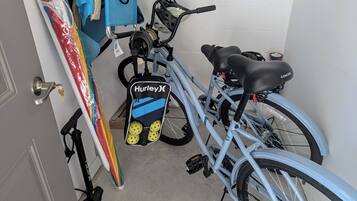 A bicycle with a Hurley brand seat, a blue bag, and a surfboard leaning against a wall.