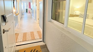 A hotel hallway with a 'hello' mat, a bed with a patterned pillow, and a window with blinds.