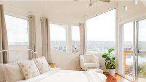 A bedroom with a large bed, a white chair, a potted plant, and a view of a cityscape through large windows. - Rhythm&Views- Hillside home in Mount Adams with Hot Tub and River Views (Cincinnati)