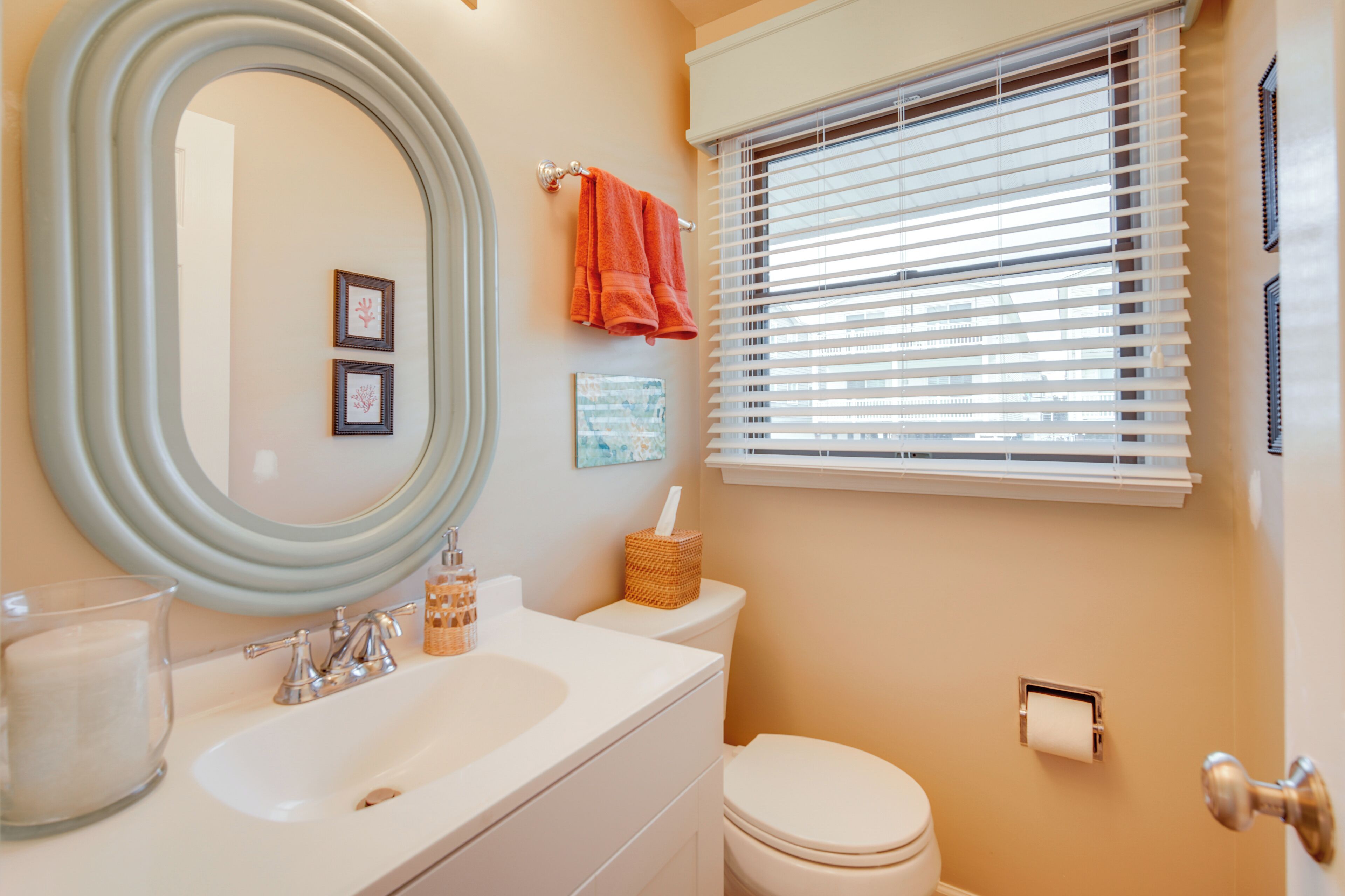 Une salle de bain avec un lavabo blanc, un grand miroir, des toilettes et une fenêtre avec des stores.