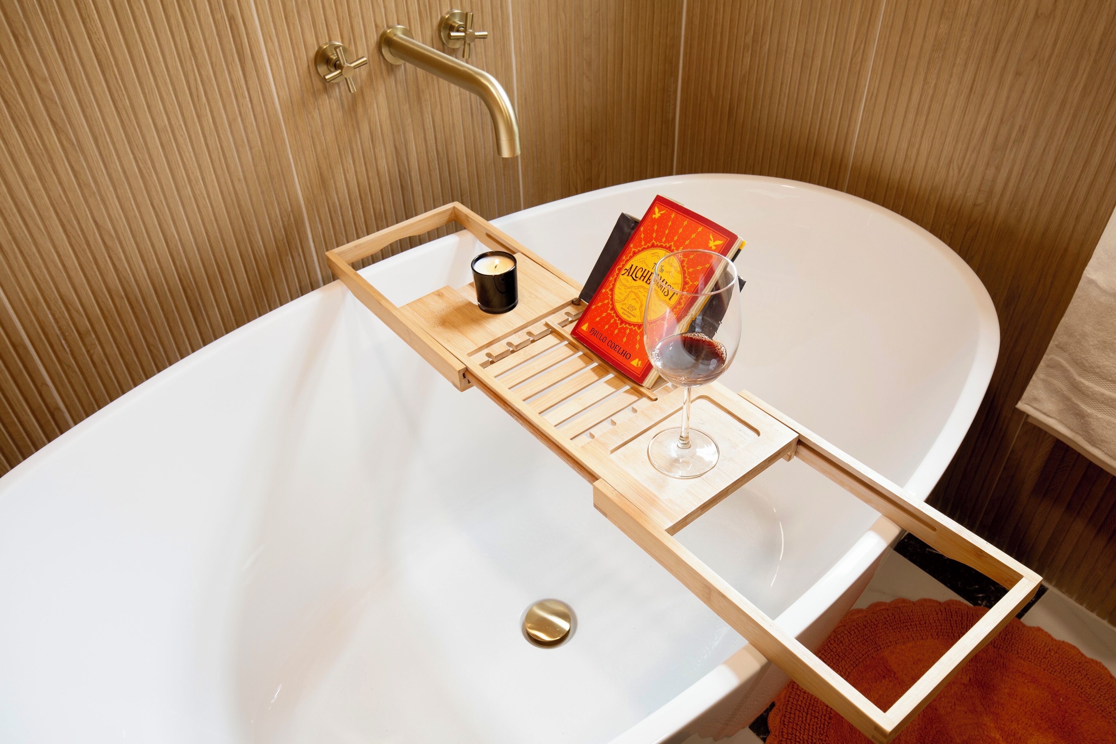 Un baño con bañera, una bandeja de madera con una vela, una copa de vino y un libro.