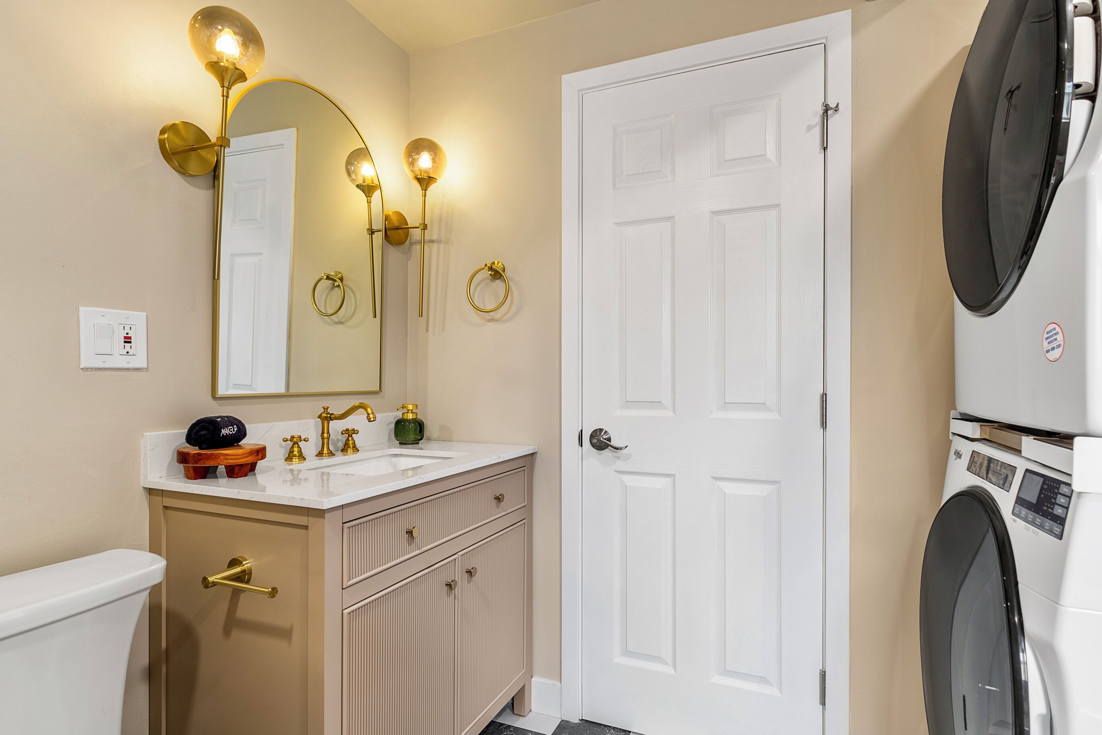 A bathroom with a toilet, sink, vanity, mirror, wall sconces, and a washing machine.