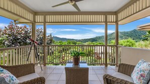 A covered balcony with wicker furniture, a telescope, and a view of lush greenery and hills. - Belle Escapes Waterfall Crossing (Redlynch)
