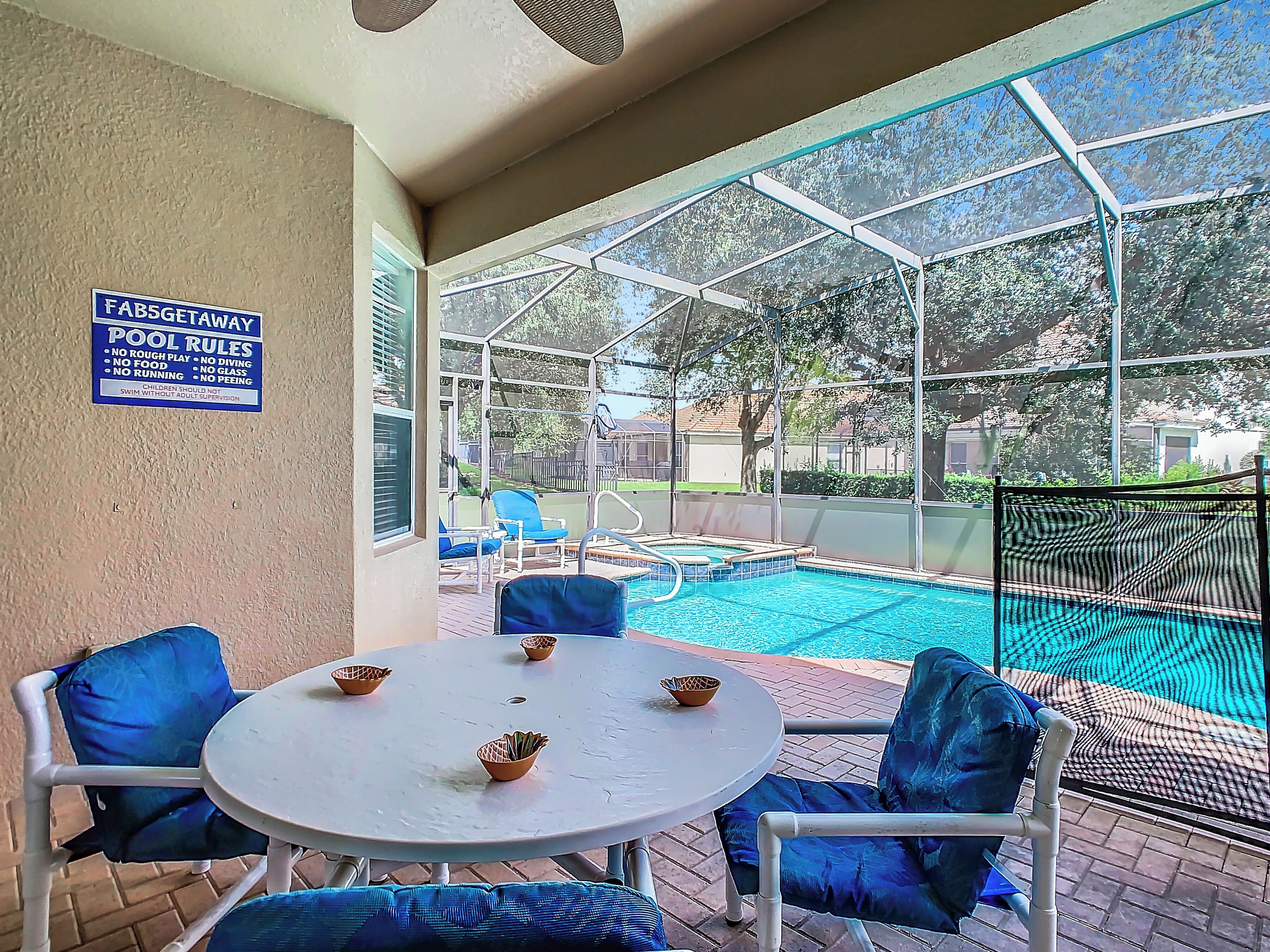 A covered outdoor pool area with a table and chairs, a sign with pool rules, and a view of the backyard.