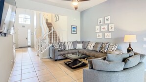 A spacious living room with a gray sectional sofa, a coffee table, and a ceiling fan. A staircase leads to an upper level, and the walls are adorned with framed artwork. - TOP-RATED Windsor Hills Resort Villa ♥ THEATER/GAME ROOM, Pool ♥ 4 Mi to Disney (Kissimmee)