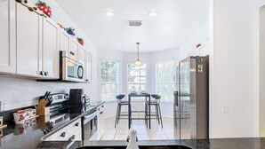 A modern kitchen with white cabinets, a stainless steel sink, a microwave, and a refrigerator. There are two bar stools and a dining table with chairs. - TOP-RATED Windsor Hills Resort Villa ♥ THEATER/GAME ROOM, Pool ♥ 4 Mi to Disney (Kissimmee)
