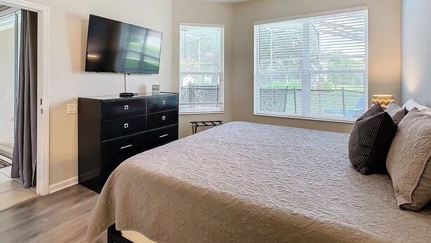 A bedroom with a bed, a TV, a dresser, and two windows with blinds.