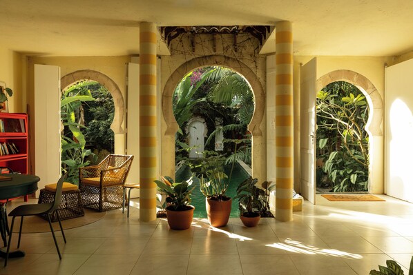 An indoor space with a tiled floor, a red bookshelf, a round table, and several potted plants. - O Portalzinho (Paraty)