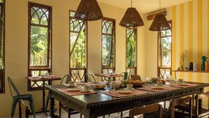 A dining area with a large wooden table set for a meal, surrounded by chairs. There are large windows with a view of greenery outside. - O Portalzinho (Paraty)