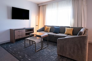 A modern hotel room with a sofa, a small table, a TV mounted on the wall, and a floor lamp.