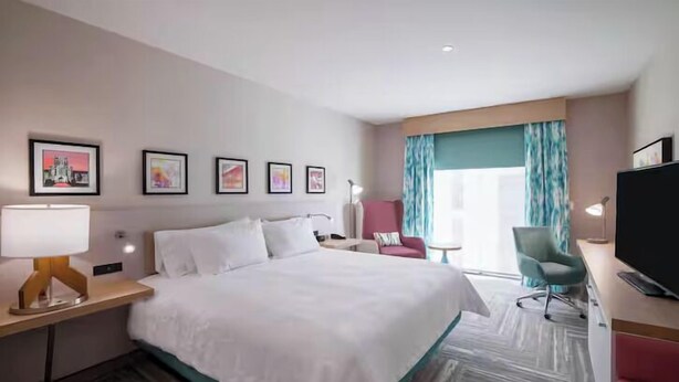 Ein Hotelzimmer mit einem großen Bett, einem Schreibtisch mit Lampe, einem Fernseher, einem Sessel und einem Fenster mit Vorhängen.