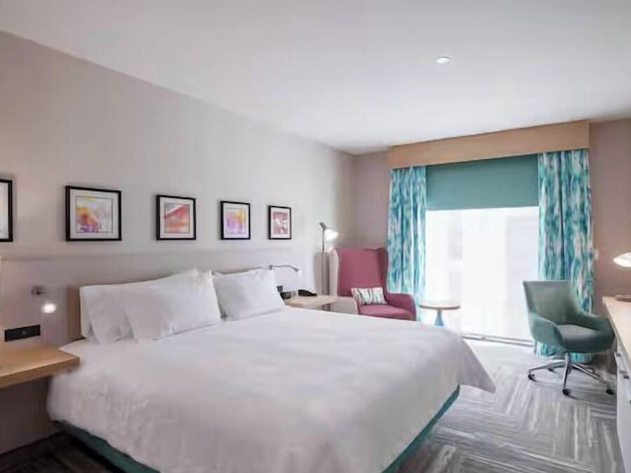 A hotel room with a large bed, a desk with a lamp, a TV, a chair, and a window with curtains.