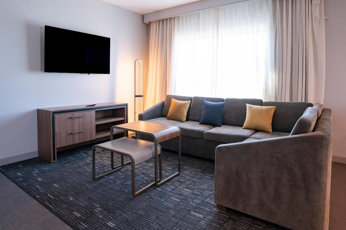 Ein modernes Wohnzimmer mit einer Couch, einem Couchtisch, einem an der Wand montierten Fernseher und einem großen Fenster mit Vorhängen.