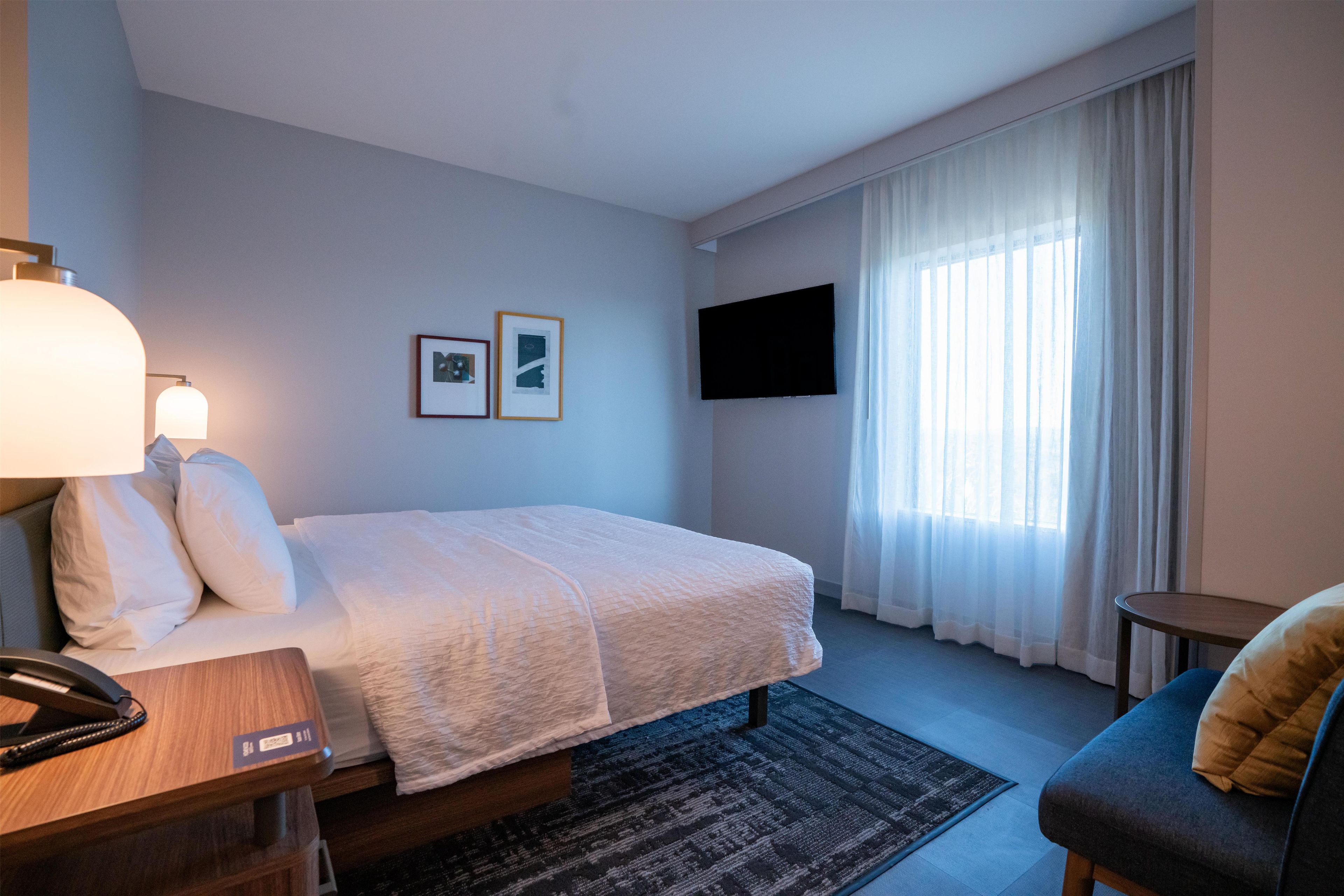 Quarto de hotel com cama, escrivaninha com telefone, cadeira, TV fixada na parede e janela com cortinas.
