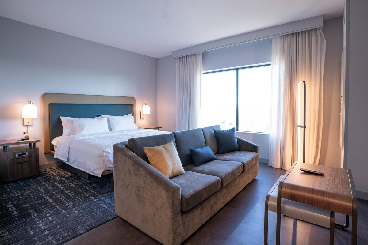 Ein modernes Hotelzimmer mit Bett, einer Sitzecke, einem Schreibtisch und einem großen Fenster mit Vorhängen.