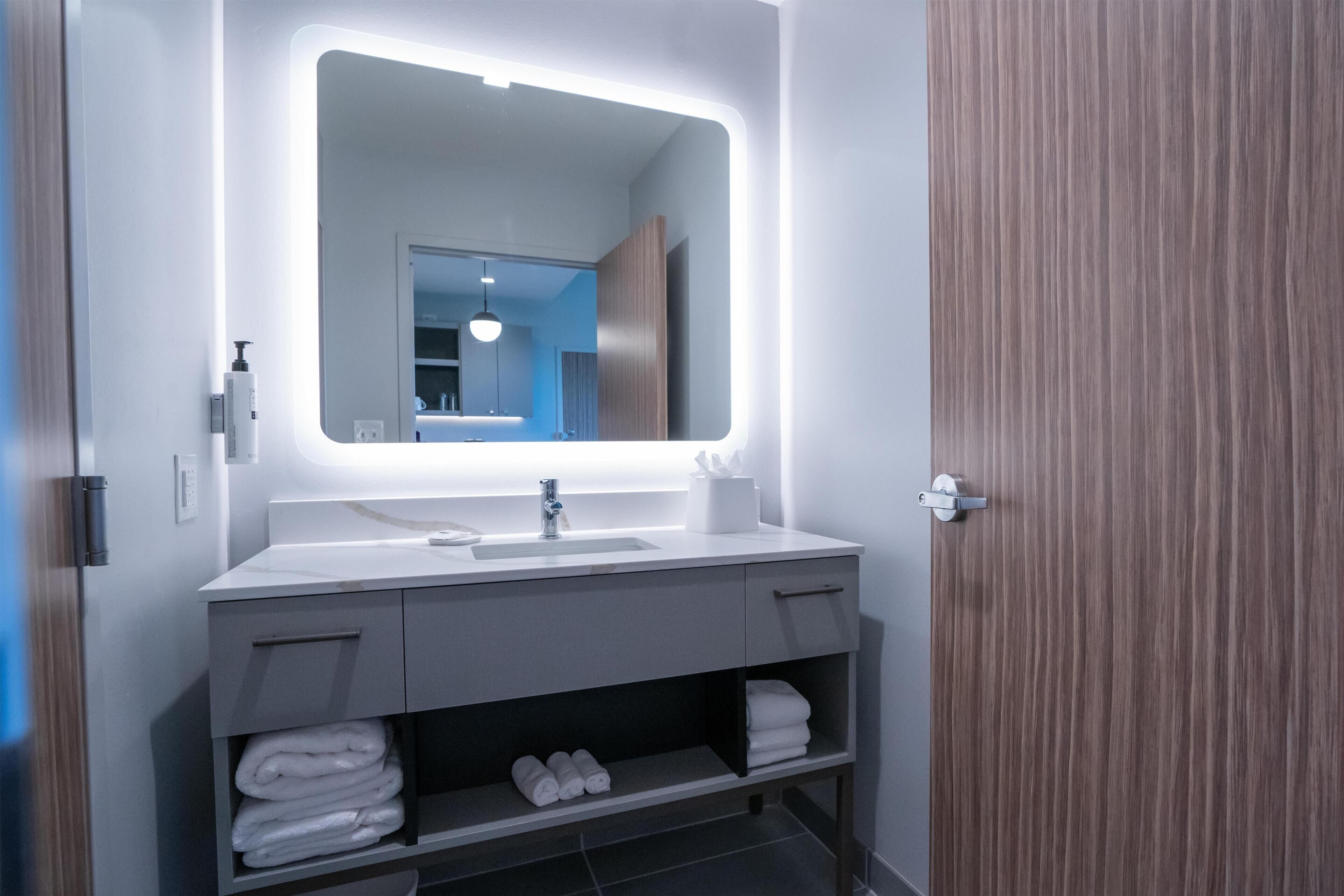 A modern bathroom with a large mirror, white vanity, and shelves for towels.